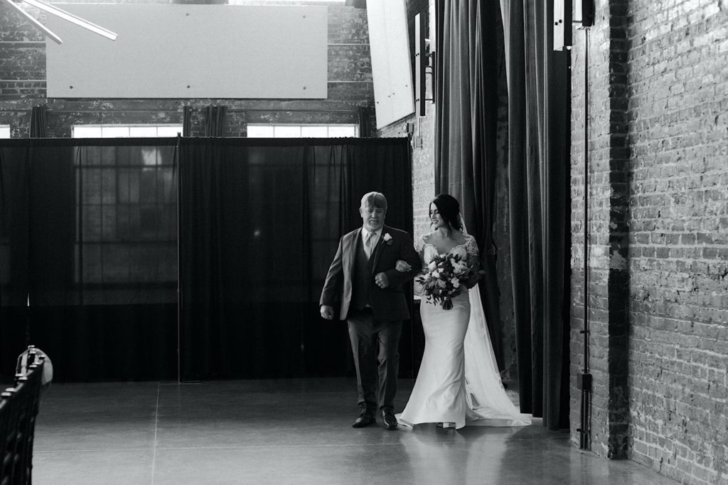 black and white photo of dad walking his daughter down the aisle on her wedding day at the cadillac service garage in greensboro north carolina